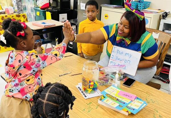 Teacher sitting at table with 3 students showing them a flash card and smiling and giving the middle student a high five