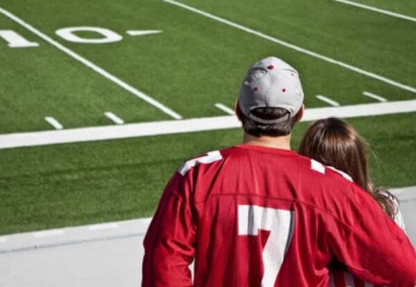 Two people with backs turned standing looking at a football field, red jerseys on.
